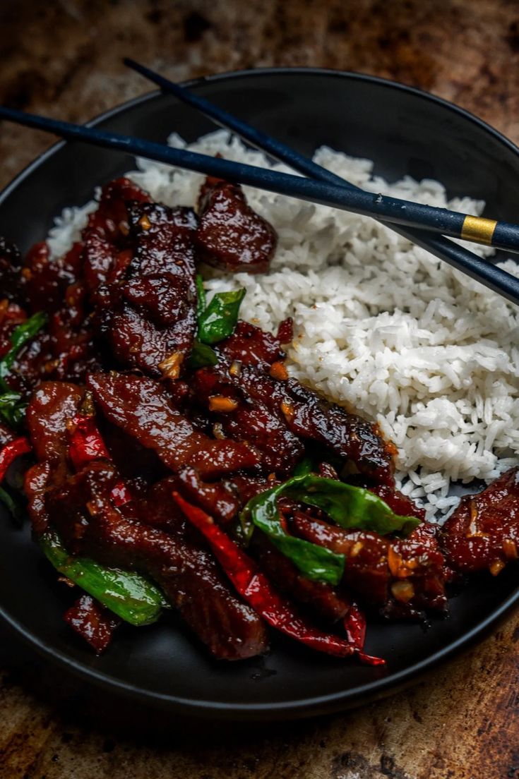 a black plate topped with rice and meat next to chopsticks on a table