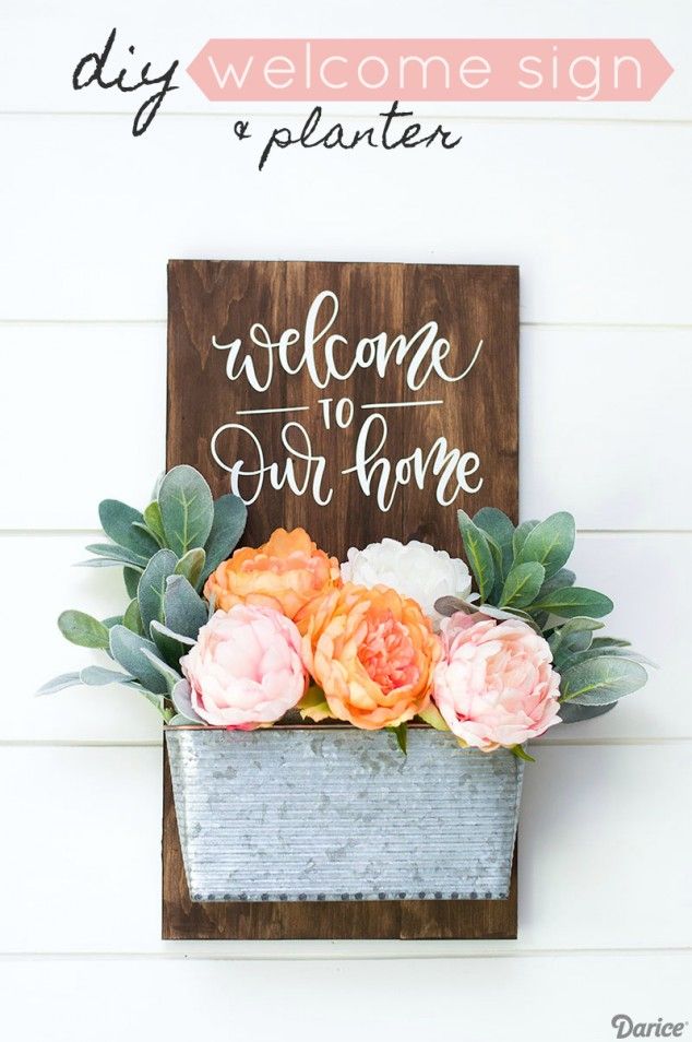 a welcome sign hanging on the side of a wall with flowers in it and greenery