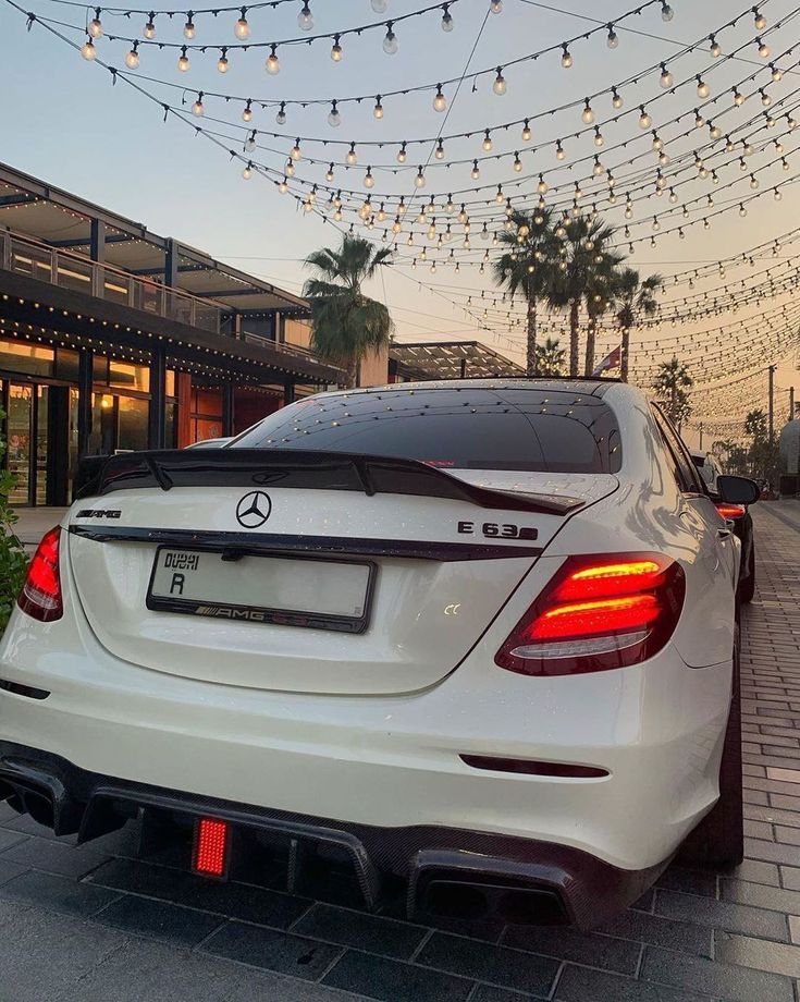 the rear end of a white car parked in front of a building with lights strung above it