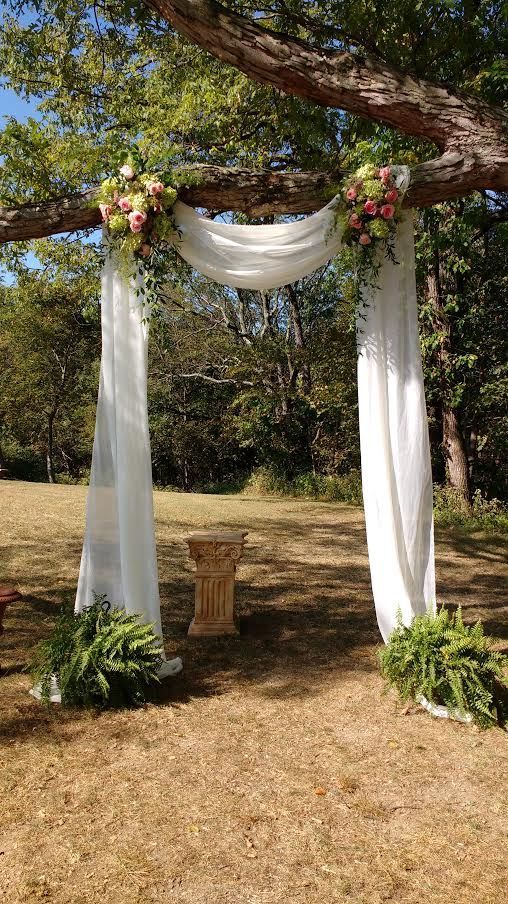 an outdoor wedding setup with white drapes and flowers on the arbor, surrounded by greenery