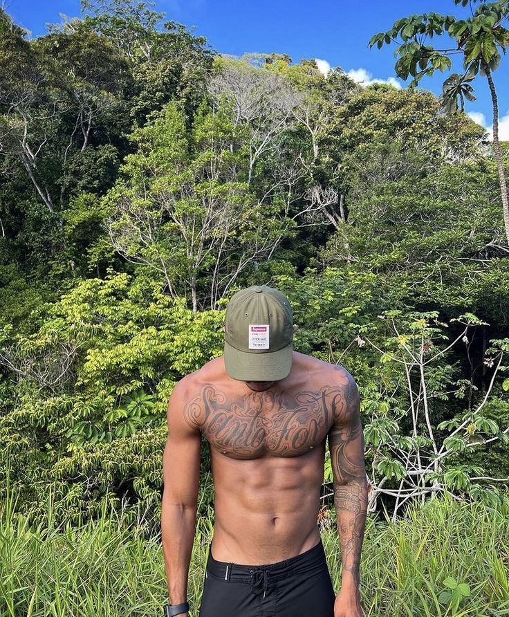 a shirtless man standing in front of some trees and bushes wearing a green hat