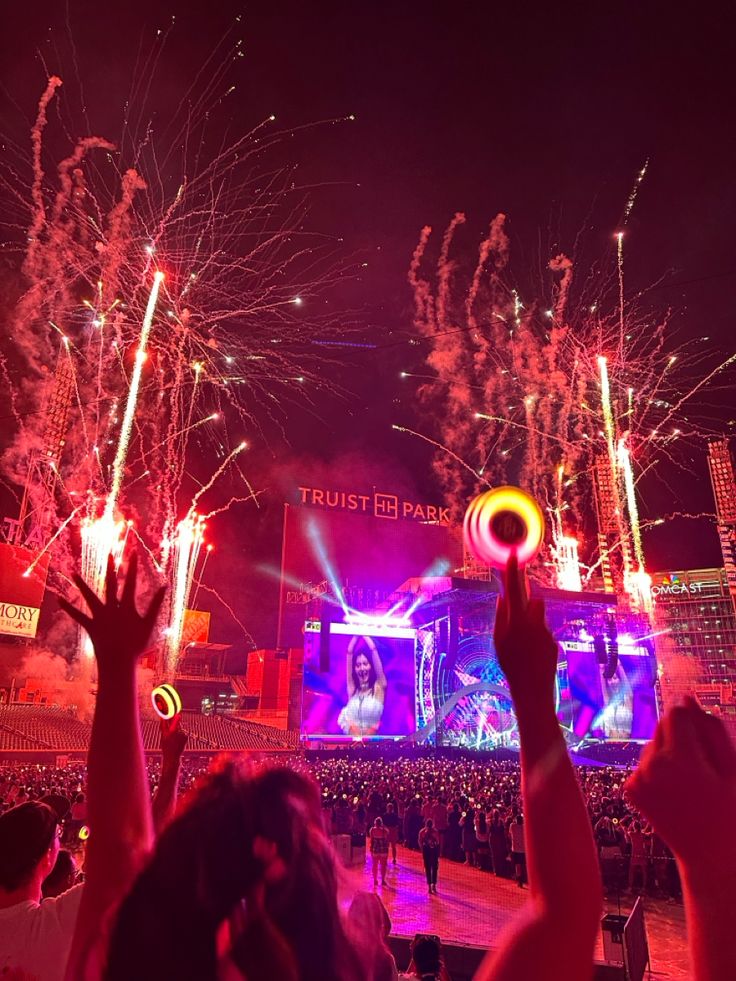 fireworks are lit up in the night sky above a crowd at an outdoor concert with people raising their hands