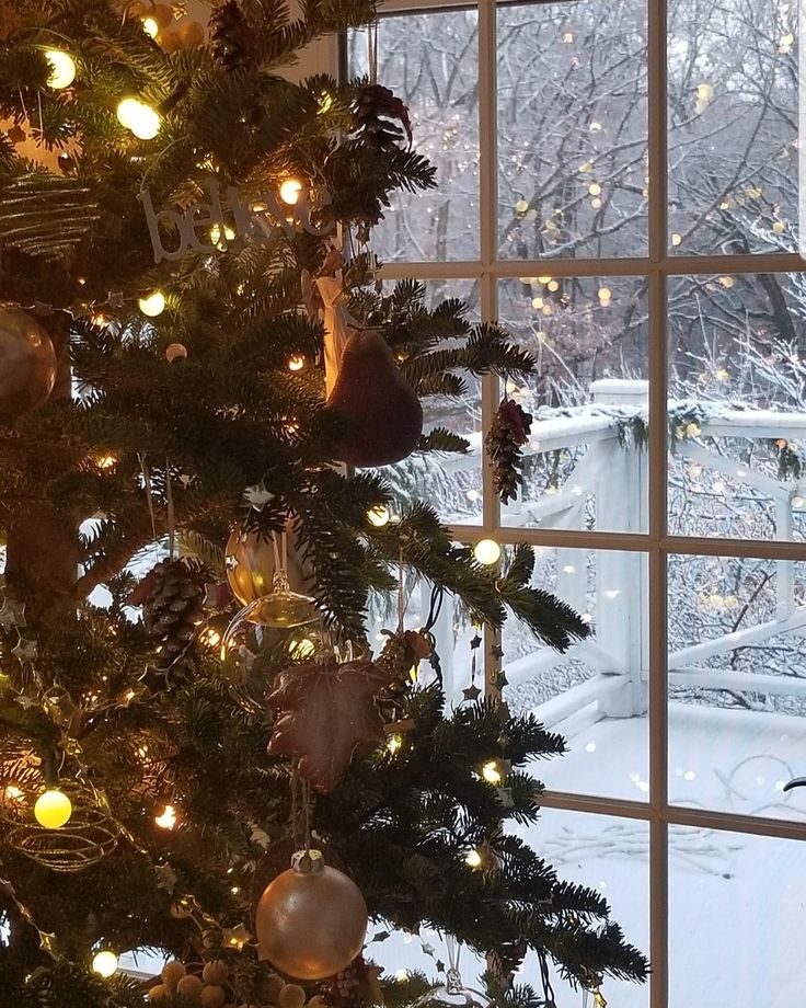 a christmas tree is lit up in front of a window with the snow outside it