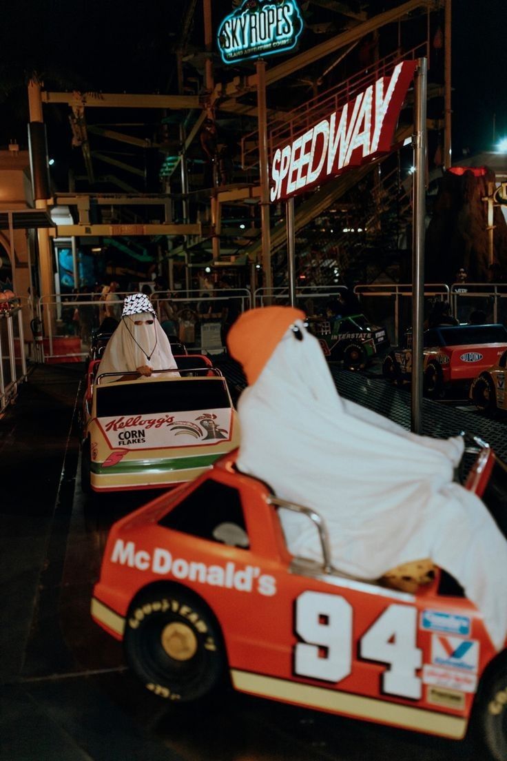 an orange race car with a white sheet on it's hood parked in front of a speedway sign