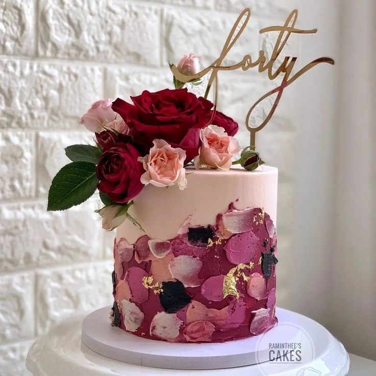 a pink and red cake with flowers on top sitting on a white table next to a brick wall