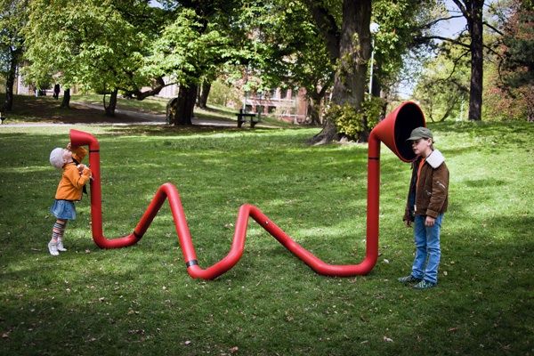 two children standing in the grass near a red tube shaped like a letter m and w