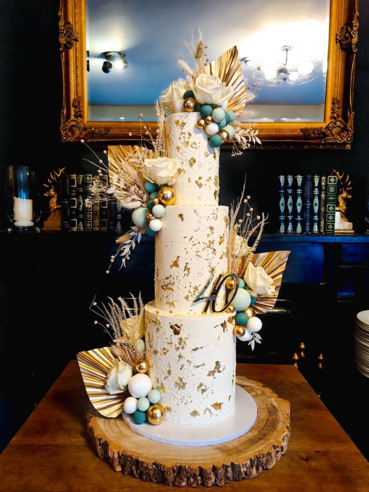 a white wedding cake decorated with gold and blue decorations on a wooden table in front of a mirror