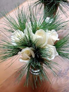 two vases with white roses and pine needles on a wooden table in front of a brick wall