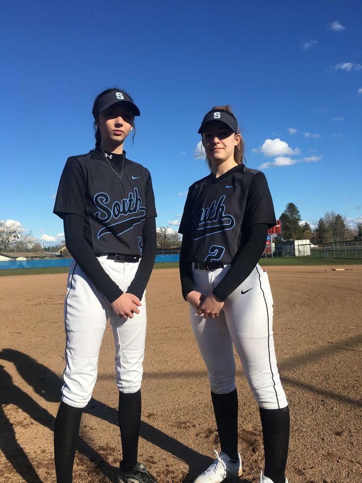 two women in baseball uniforms standing next to each other on a field with dirt and grass