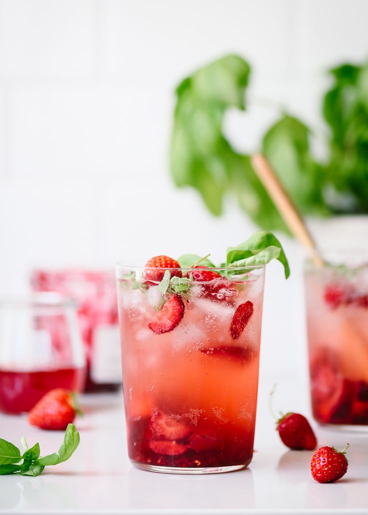 two glasses filled with liquid and strawberries next to each other on a white table