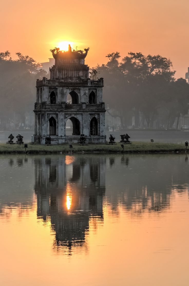 the sun is setting behind an old building on the water's edge in front of trees