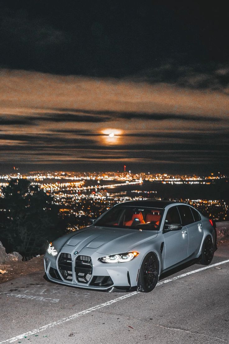 a silver sports car parked on the side of a road with city lights in the background