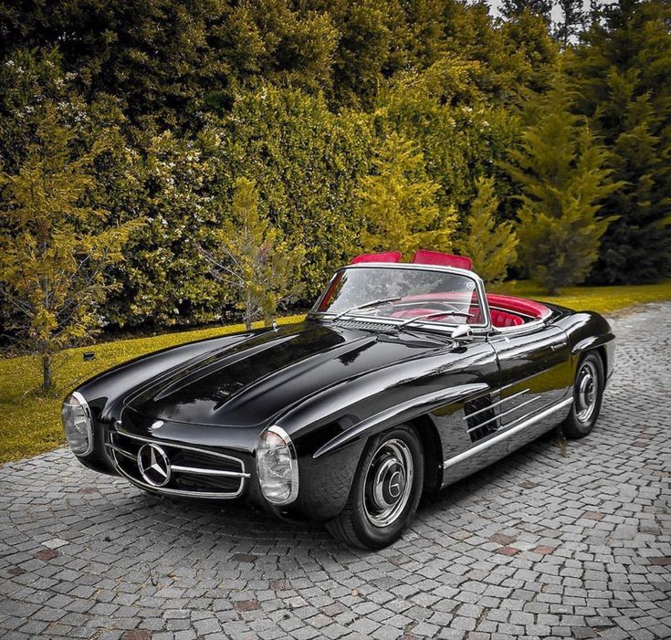 an old black mercedes sports car parked in front of some trees and bushes on a cobblestone driveway