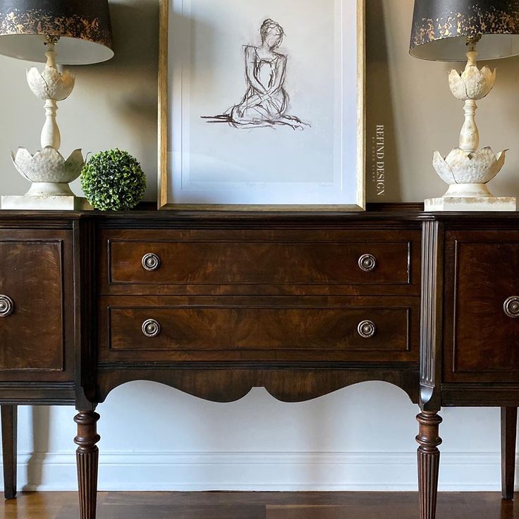 two lamps are sitting on top of an antique dresser with a drawing in the background