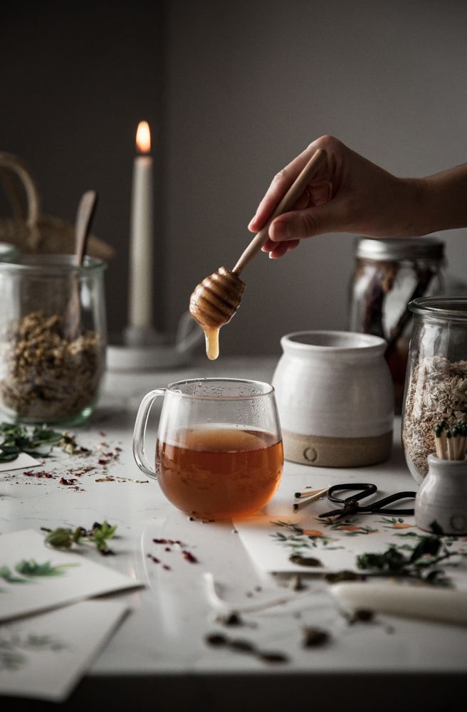 a person holding a wooden spoon over a cup filled with tea next to jars and candles