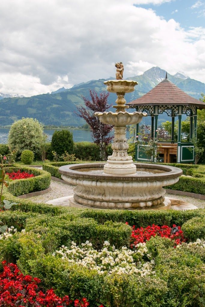 a fountain in the middle of a garden surrounded by flowers and shrubs with mountains in the background