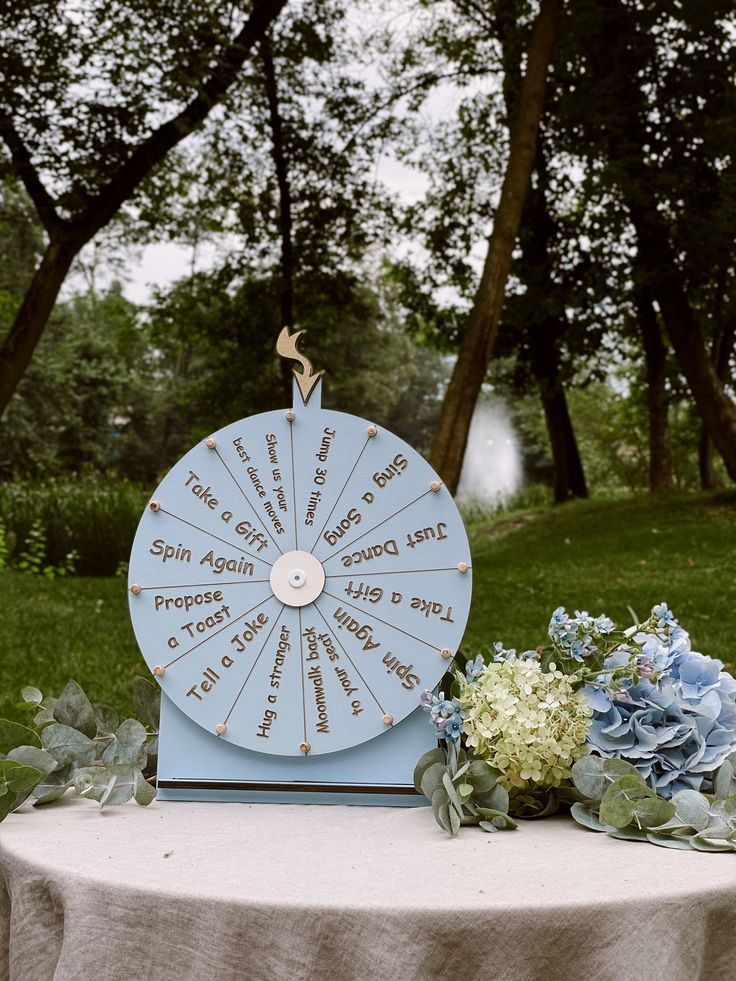 a table topped with a blue wheel of fortune next to flowers and greenery in front of trees