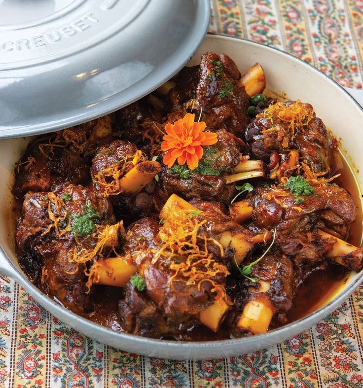 a bowl filled with meat and vegetables on top of a colorful table cloth next to a pot