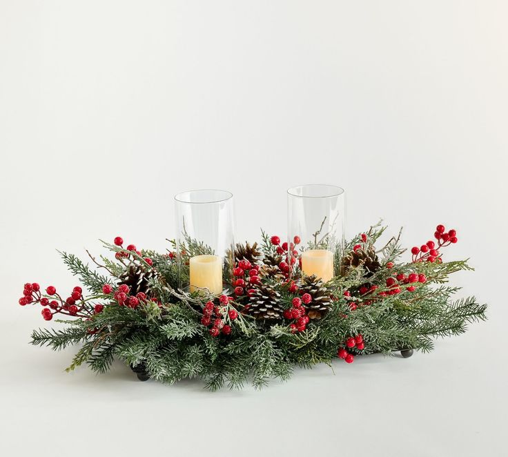 two votive candles are sitting on a christmas centerpiece with pine cones and red berries