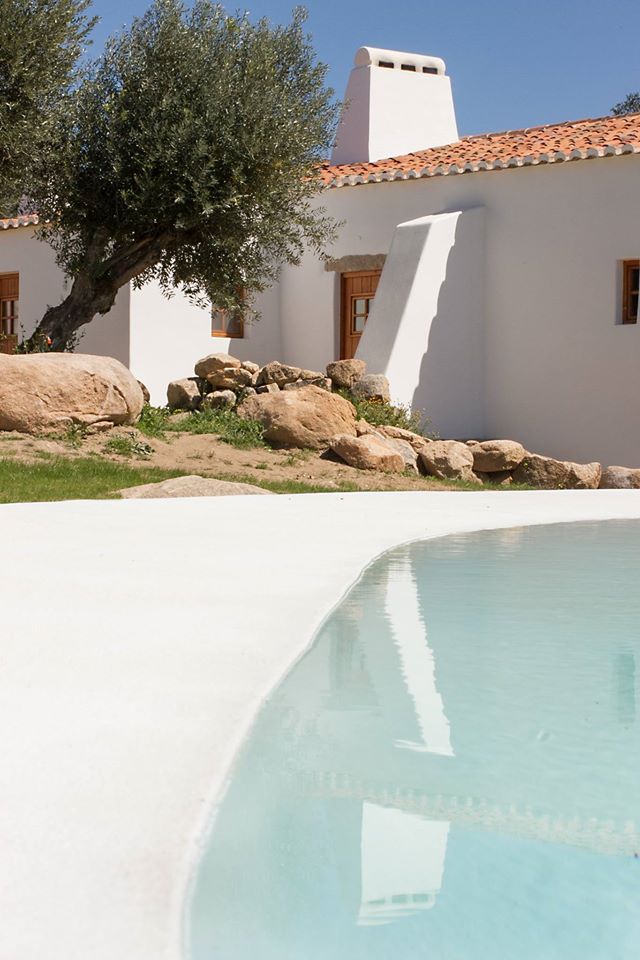 an outdoor swimming pool in front of a white house with trees and rocks around it