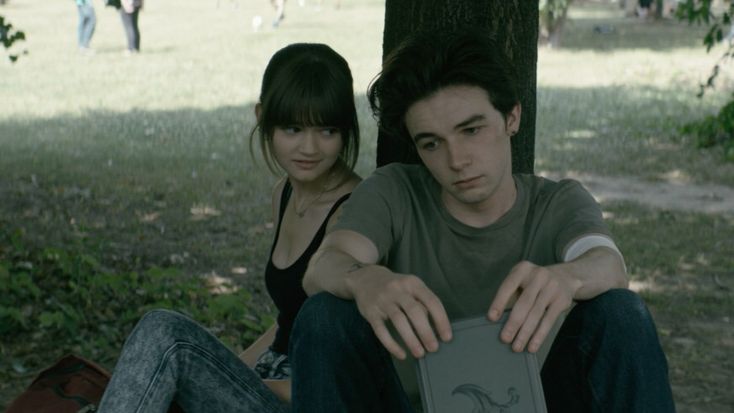 two young people sitting under a tree in the park, one holding an open book