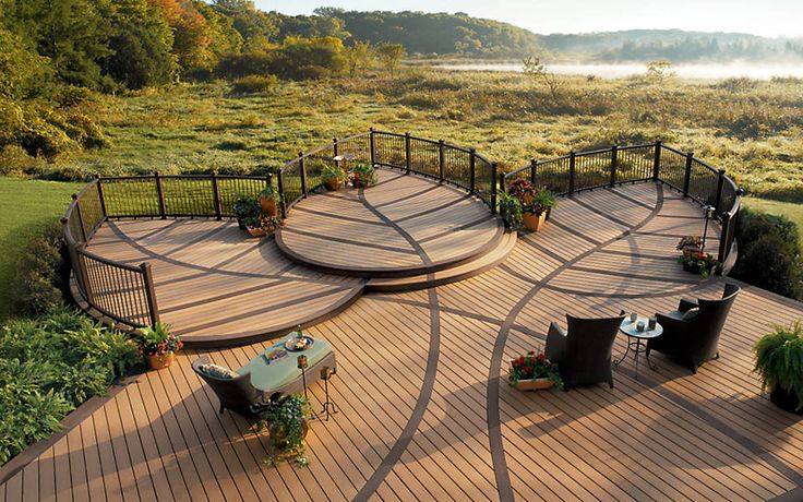 a wooden deck with chairs and tables in the middle of an open field, surrounded by greenery