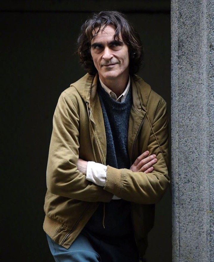 a man leaning against a wall with his arms crossed and looking at the camera while wearing a brown jacket