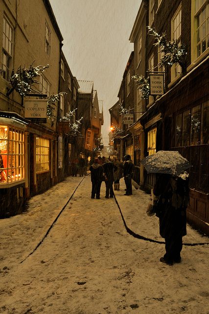 people walking down an alley way in the snow with umbrellas and lights on buildings