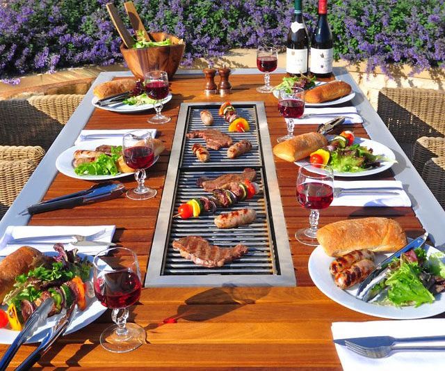 an outdoor grill with food and wine on the table outside in front of purple flowers