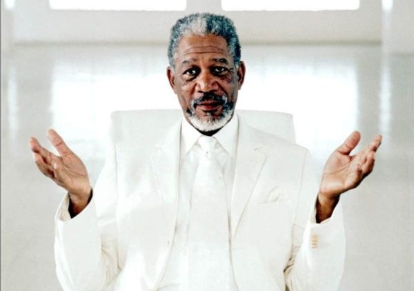 an older man in a white suit and tie sitting on a chair with his hands out