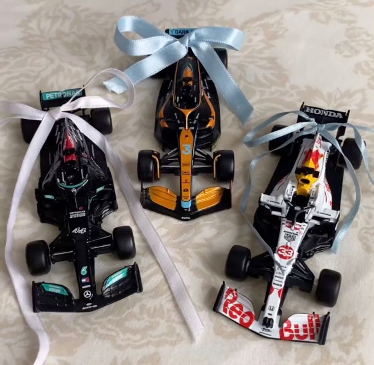 three toy race cars with ribbons tied around them on a bed sheet, one is black and the other is white