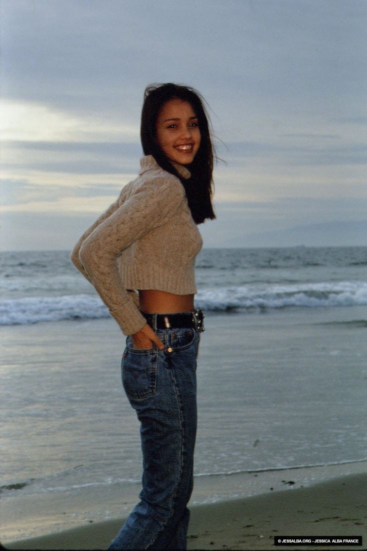 a woman standing on the beach with her hands in her pockets and smiling at the camera