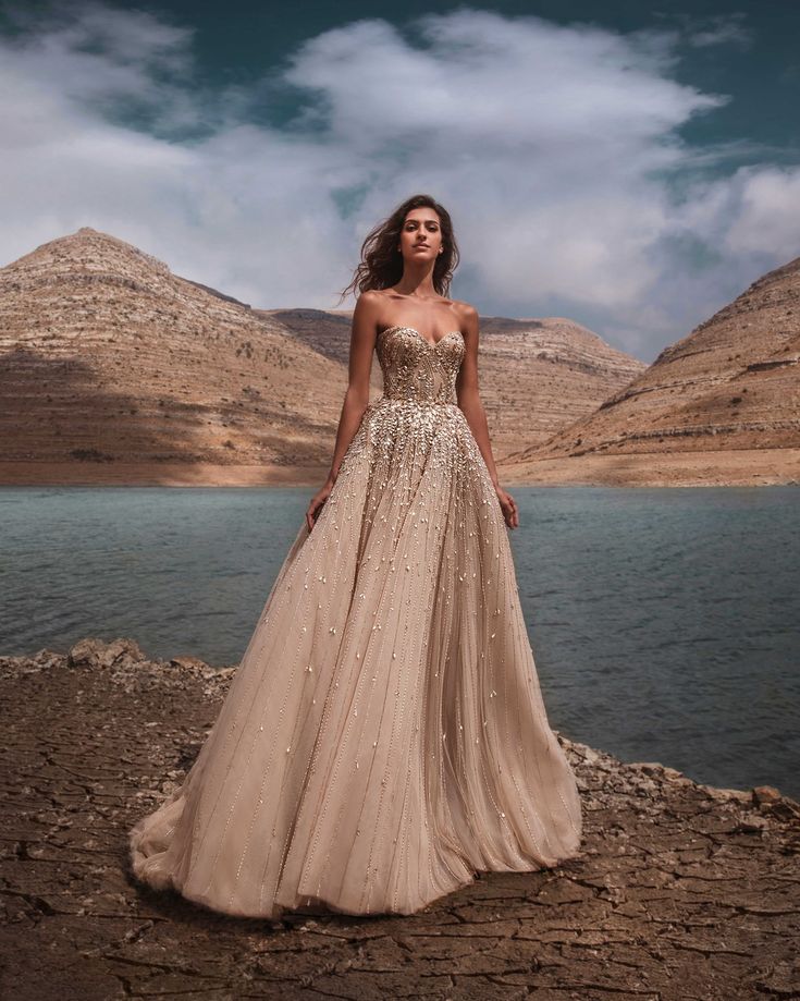 a woman in a long dress standing on rocks near the water with mountains behind her