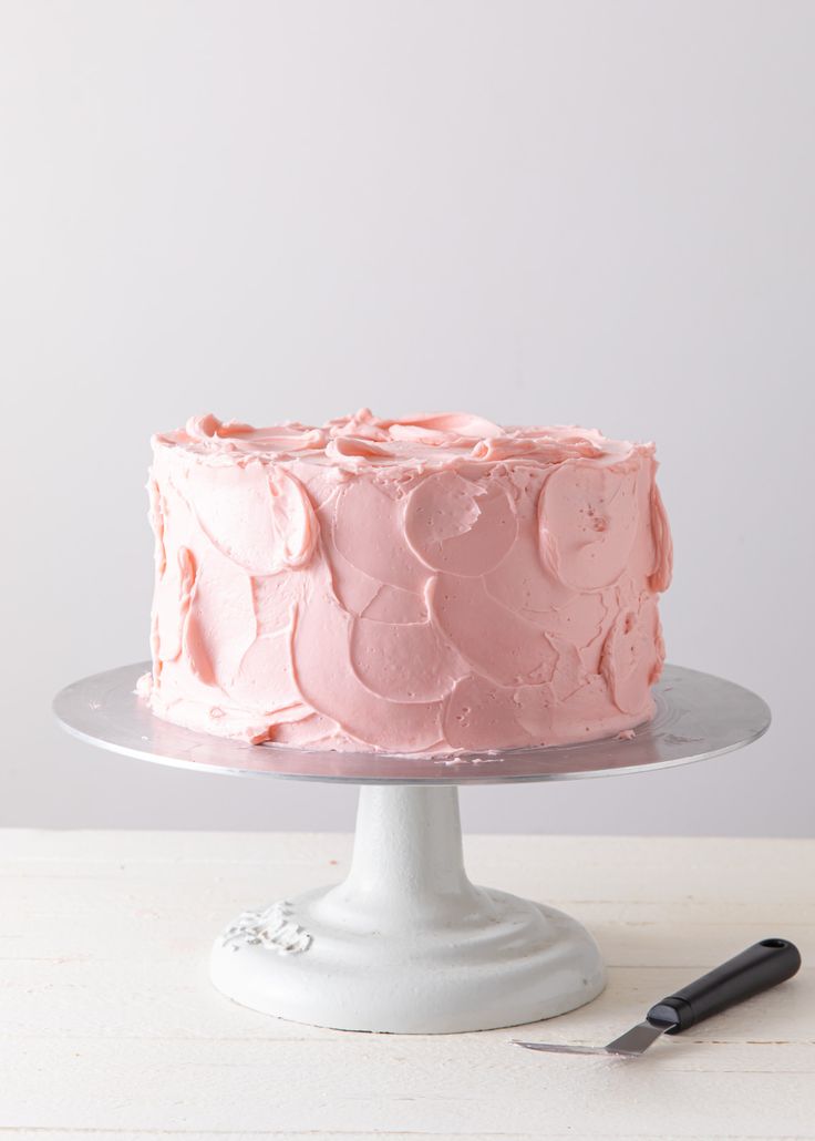 a pink frosted cake sitting on top of a white cake plate next to a knife