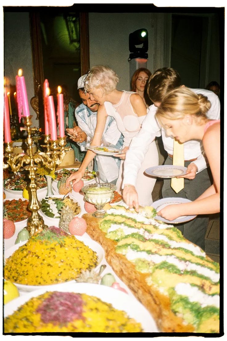 several people standing around a table with food on it and candles burning in the background