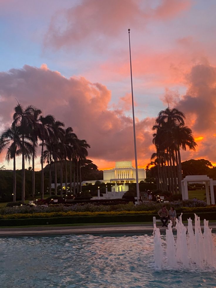 a fountain in front of palm trees and a building with a clock tower at sunset