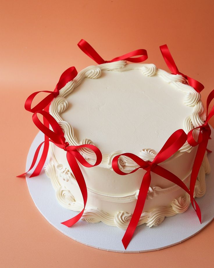 a white cake with red ribbon on top is sitting on a pink surface and it looks delicious