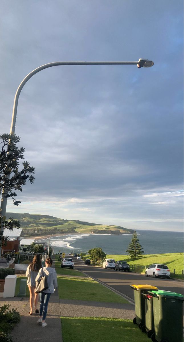 two people walking down a sidewalk next to a street light with the ocean in the background