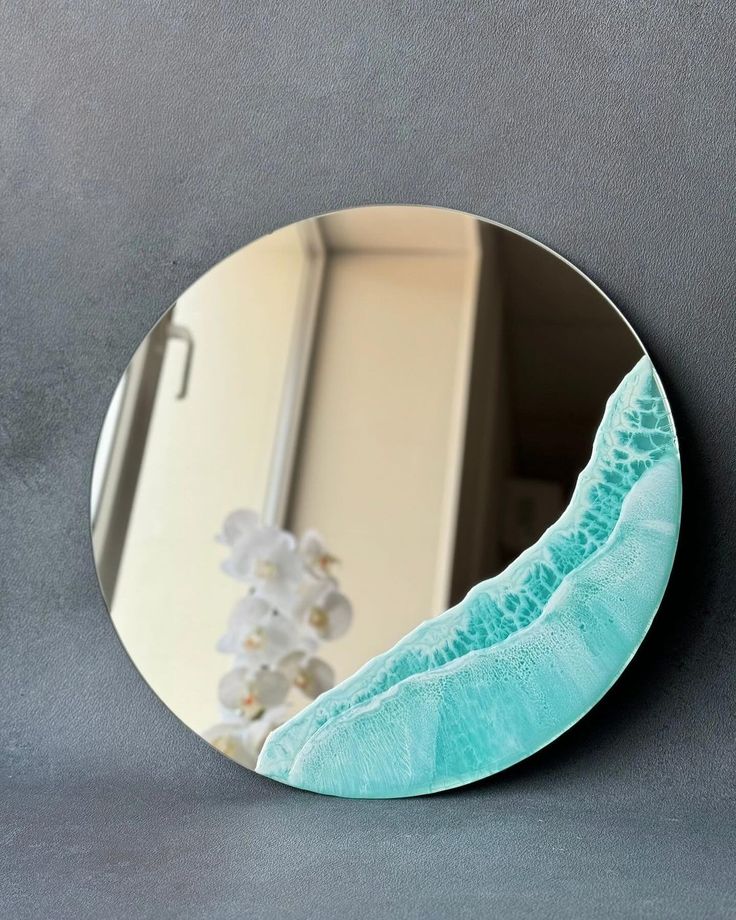 a round mirror on the wall above a vase with flowers in it and a blue wave