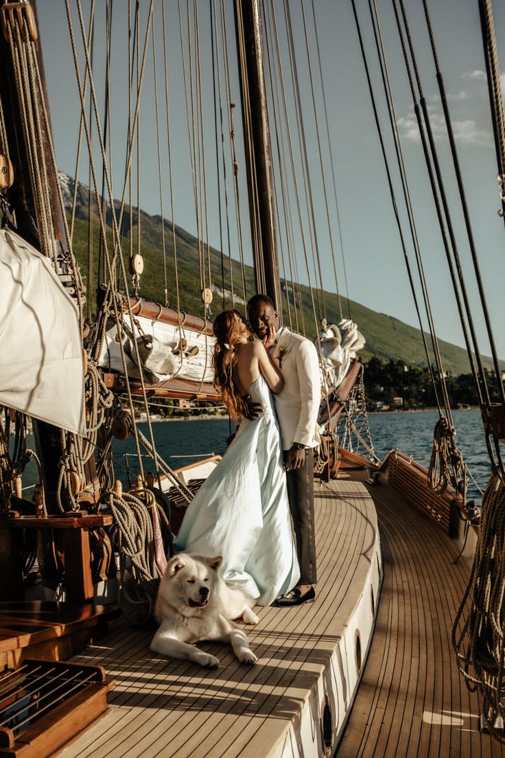 a bride and groom standing on the deck of a sailboat with their white dog