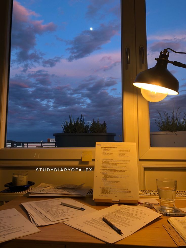 an office desk with papers on it and a lamp in front of the window at dusk