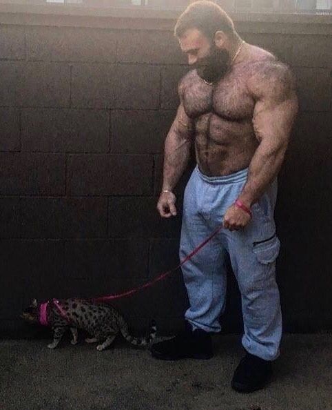 a man with a beard standing next to a cat on a leash and wearing sweatpants