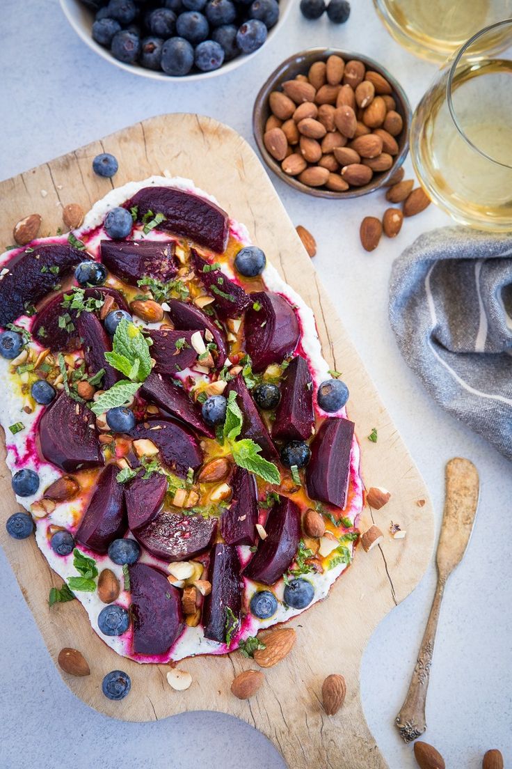 a wooden cutting board topped with beets and blueberries next to bowls of nuts