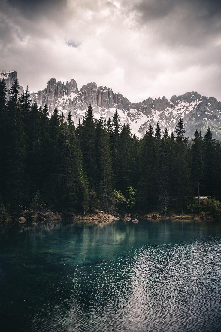 a lake surrounded by trees and mountains under a cloudy sky with snow on the top