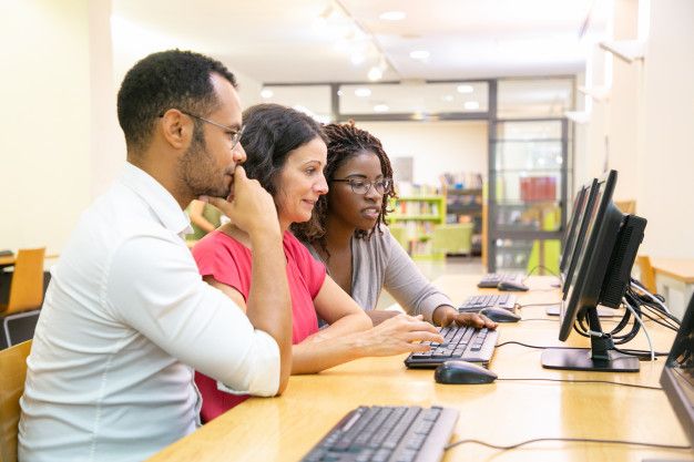 three people sitting at a table with computers in front of them, looking at the screen