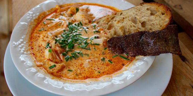 a white plate topped with soup and meat on top of a wooden table next to bread