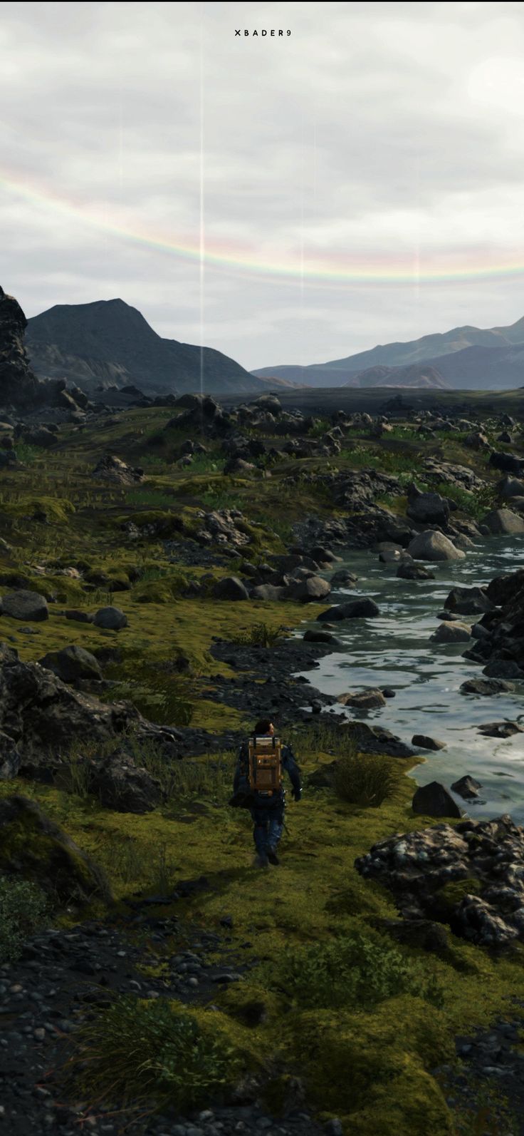 a man walking across a lush green field next to a river with a rainbow in the sky