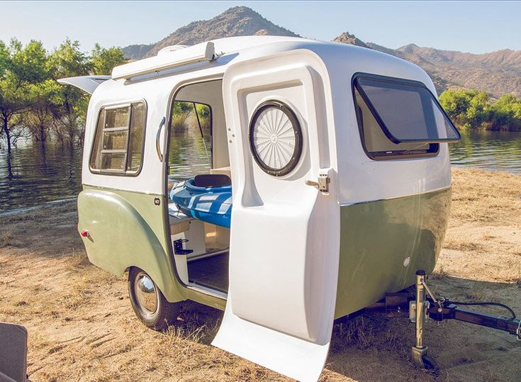 an old camper trailer is parked on the sand near water and trees with mountains in the background