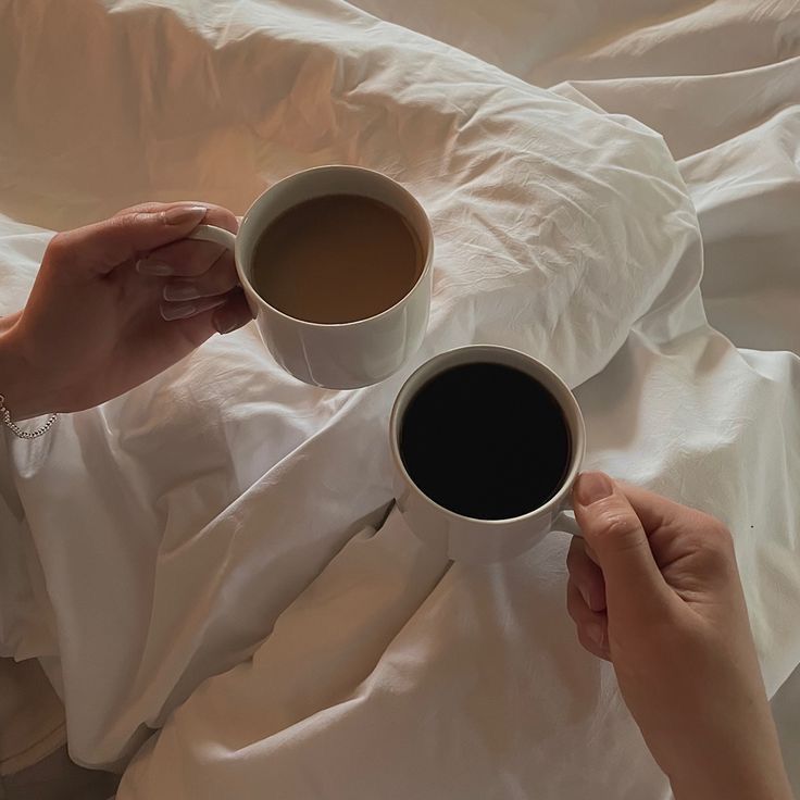 two hands holding cups of coffee on top of a white sheet covered bedspread