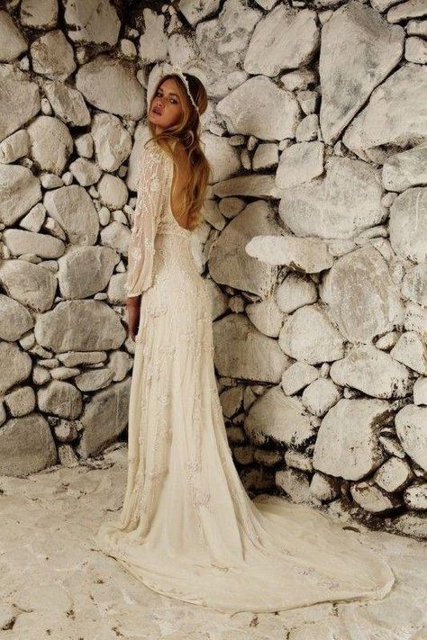 a woman standing in front of a stone wall wearing a wedding dress with long sleeves